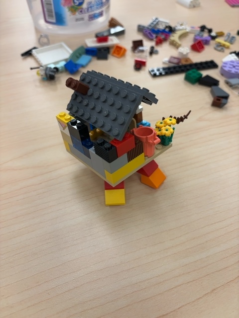 A Fast Brix! gallery build by @RidgedBrick for the #HouseChasingFeet prompt: a colorful LEGO house with a gray sloped roof, chimney, window and flowers, standing on big chunky red, yellow and orange feet.
